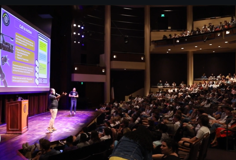 Two speakers on stage presenting to a packed high school auditorium